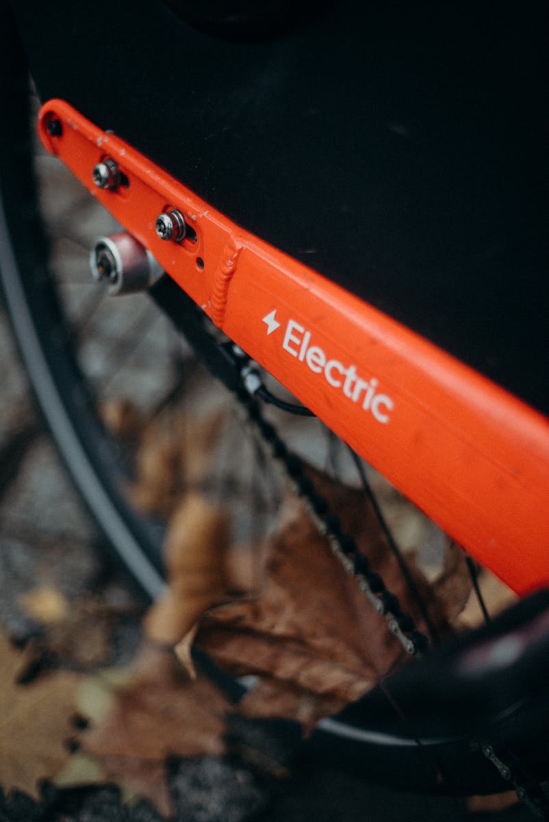 A detailed close-up of a bright orange electric bicycle with autumn leaves scattered around on a city street.